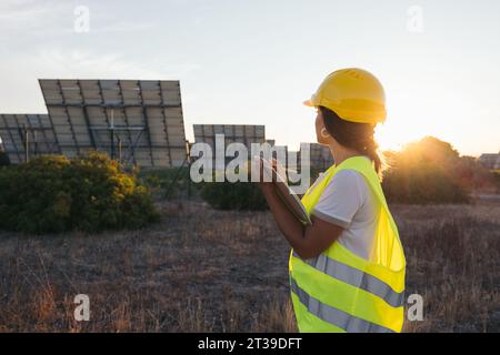 From behind a technical woman taking data next to some solar panels with a vest and protective helmet Stock Photo