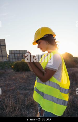 From behind a technical woman taking data next to some solar panels with a vest and protective helmet Stock Photo