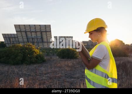 From behind a technical woman taking data next to some solar panels with a vest and protective helmet Stock Photo