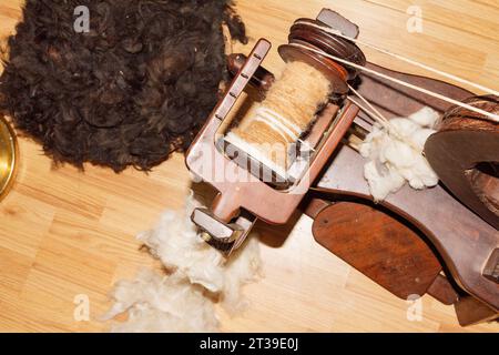 A vintage wooden spinning wheel with a spool of light brown yarn, set against a pile of raw dark brown wool on a wooden floor. Stock Photo
