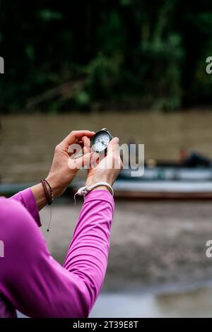 Back view of crop anonymous female traveler in casual clothes holding vintage compass in hands against blurred background of people in boats in Amazon Stock Photo