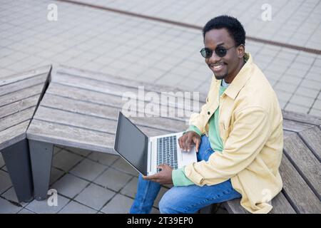 From above of smiling young African American male in sunglasses looking at camera while sitting on bench with laptop in daylight Stock Photo