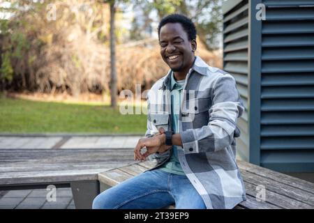 Smiling young African American male eyes closed while sitting on bench near blurred building with sidewalk and holding watch in daylight Stock Photo