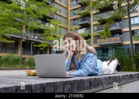 Happy blond haired student woman with braces lying in wooden bench and looking at laptop in lunchtime outdoors. Stock Photo