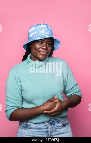 An African woman in a blue cloud-patterned bucket hat and turquoise turtleneck sweater, posing against a pink background Stock Photo