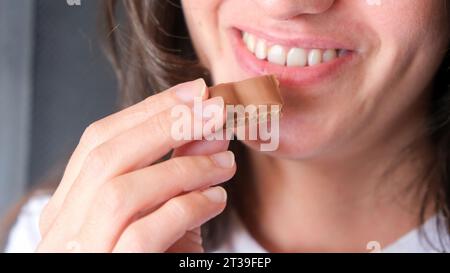 A young female standing in front of a white background, holding a box of chocolates in her left hand and a piece of chocolate in her right hand Stock Photo