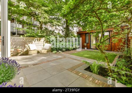 Backyard area with chairs on tiled patio and small garden by wooden shed surrounded with lush trees and plants Stock Photo