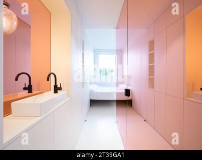 A stylish bathroom area with dual sinks against a backdrop of gradient colored walls transitioning from pink to orange, leading to a bedroom view. Stock Photo