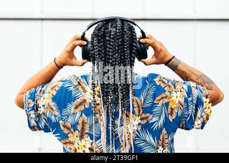 Latin man from behind of a latin man with detailed braids listening music with his black headphones, adorned with tattoos and wearing a floral blue sh Stock Photo