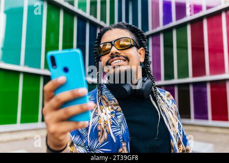 Latin man with braids holding a blue phone, wearing headphones and sunglasses, against colorful windows. Stock Photo