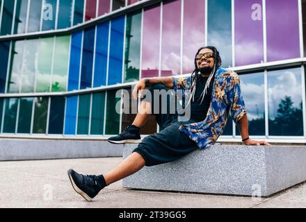 Happy latin man with braids and sunglasses sitting relaxed in front of a vibrant glass building. Stock Photo