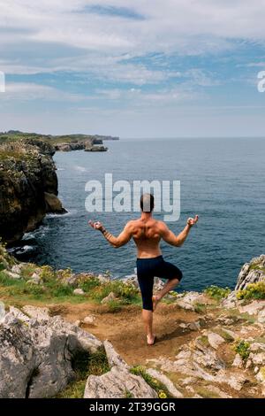 Mature man practicing yoga with a tranquil ocean backdrop, balancing on one foot while extending his arms in a scenic rocky location. Stock Photo
