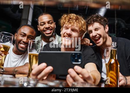 positive interracial men taking selfie on smartphone and holding beer during bachelor party in bar Stock Photo
