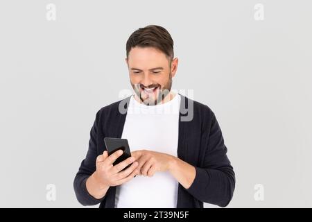 Happy young  bearded man using mobile smart phone for mobile application online, social media, on gray background. Delighted happy man expressing surp Stock Photo