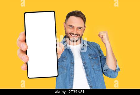 Handsome young man holding a smartphone mockup with a blank screen and smiling on an orange background. Celebrating success. Gadget with empty space f Stock Photo