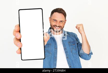 Handsome young man holding a smartphone mockup with a blank screen and smiling on a white background. Celebrating success. Gadget with empty space for Stock Photo