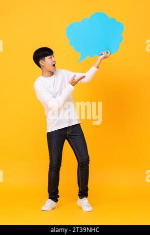 Full length portrait of young Asian man standing and looking sideways to speech bubble in isolated yellow color studio background Stock Photo