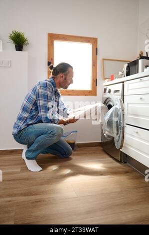 Young adult handsome Caucasian man doing the laundry at home, taking off laundered clothing out of a washing machine. Washing clothes. The concept of Stock Photo