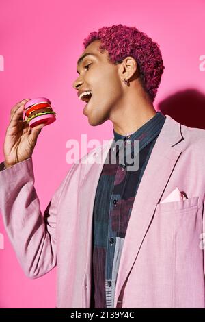 cheerful young man in pink blazer with silver earrings eating mini burger posing on pink backdrop Stock Photo