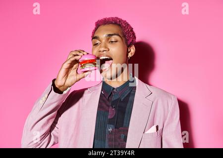 stylish african american man with silver earrings eating mini burger posing on pink backdrop Stock Photo
