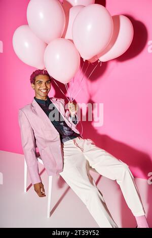 joyous man sitting unnaturally on white chair with balloons and smiling weirdly, acting like doll Stock Photo