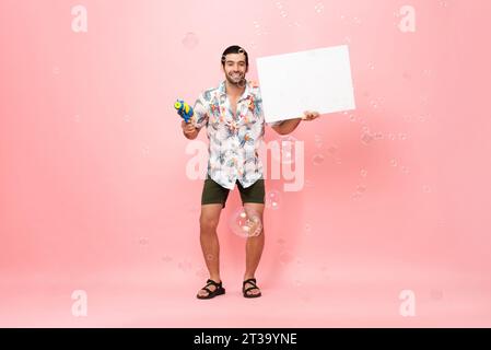 Handsome smiling young Caucasian tourist man holding water gun and empty white board with space for text in studio pink background for Songkran festiv Stock Photo