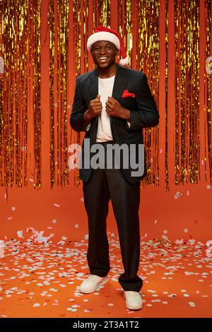 cheerful african american man in santa hat and black suit on orange backdrop with shiny decor Stock Photo