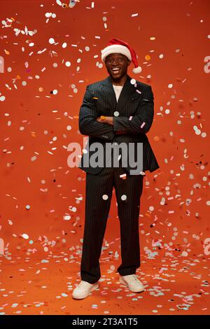 excited african american man in black suit and santa hat smiling under confetti on red backdrop Stock Photo