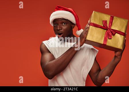 surprised african american man in santa hat holding gift box and looking at camera on red backdrop Stock Photo
