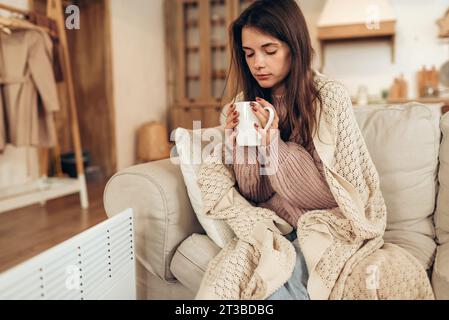 Woman freezes in winter time. Young woman with plaid near electric heater, holding a cup of hot drink Stock Photo