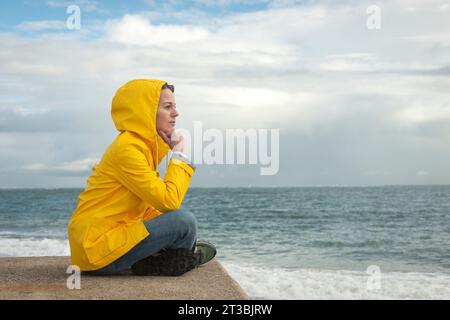 woman wearing a yellow hooded coat sitting looking at the sea, deep in thought. alone. Stock Photo