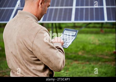 Professional investor calculating his possible profit from alternative enegry sources. Man using calculator to count exact sum of income. Back view of man counting on background of solar panels. Stock Photo