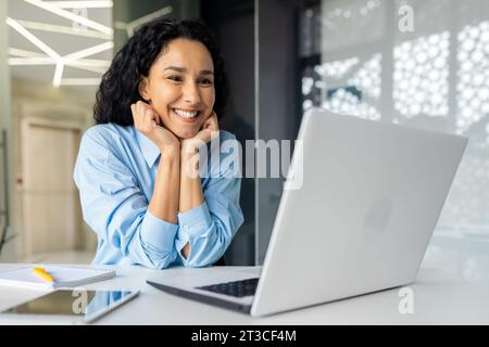 Happy and successful businesswoman reading from laptop screen, satisfied with achievement results hispanic woman dreamy thinking, sitting inside office at workplace, female financier with curly hair. Stock Photo