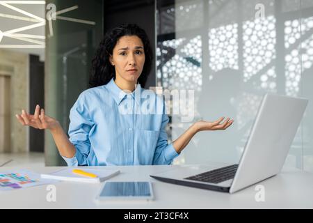 Portrait of upset disappointed indian business woman, female worker looking at camera unsatisfied, working at workplace inside office, businesswoman in shirt working with laptop. Stock Photo