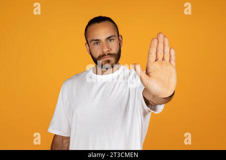 Focused bearded serious man in white t-shirt, make stop sign with his outstretched hand Stock Photo