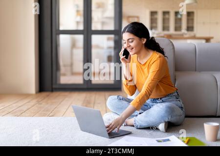 Home Office. Smiling Young Indian Woman Using Laptop And Talking On Cellphone Stock Photo