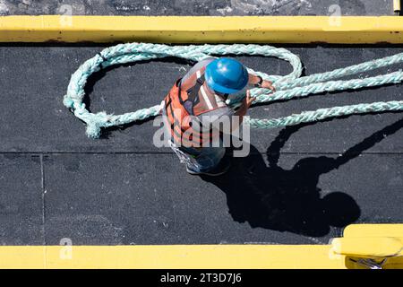 Overhead shot of a worker wearing a reflective vest and blue helmet, pulling a thick rope on a ship. Stock Photo