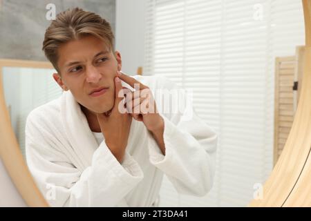 Upset young man looking at mirror and popping pimple on his face indoors. Acne problem Stock Photo