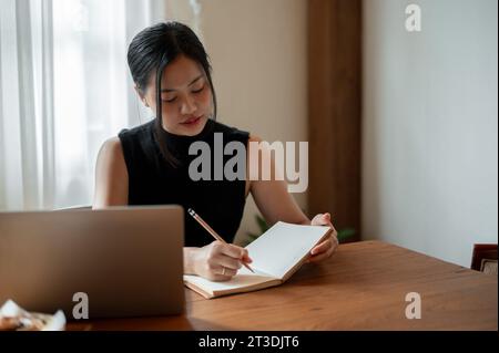 A beautiful millennial Asian woman working remotely at a coffee shop is making a list or writing some ideas down in her book. Stock Photo