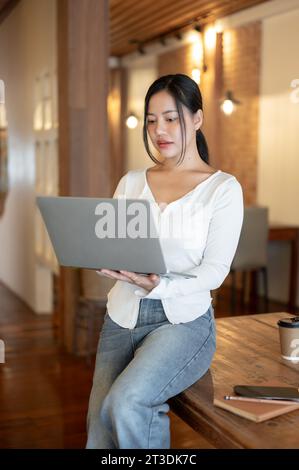 A gorgeous and focused millennial Asian woman is leaning on a table and working on her laptop in a coffee shop co-working space. Stock Photo