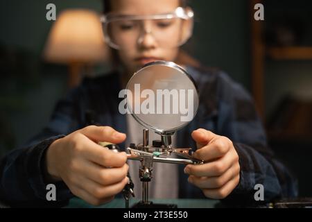 Teenager girl with safety glasses, future electronics technician, learning how to solder printed circuits, looking through a magnifying glass Stock Photo