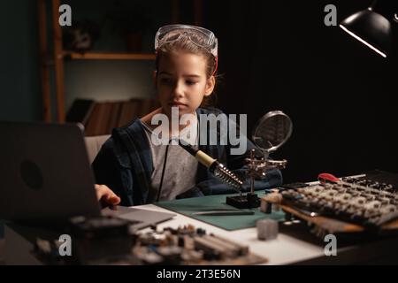Teen girl learns electronics and soldering wires on an old circuit board. Child works on a motherboard in her room. teenager is wearing safety goggles Stock Photo