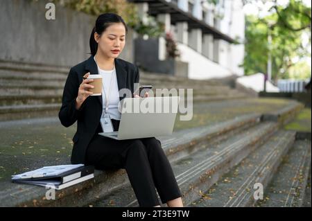 A busy and concentrated millennial Asian businesswoman is sipping coffee and responding to messages on her smartphone while working on her laptop on t Stock Photo