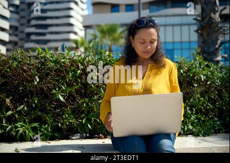 African American adult woman working remotely on laptop, typing text, sitting on a stone bench outdoor. Online business and freelance occupation conce Stock Photo
