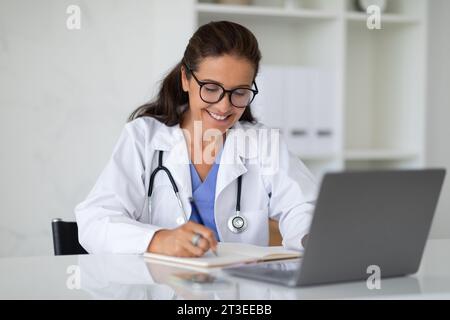 Mature woman doctor sitting at desk with laptop, taking notes Stock Photo
