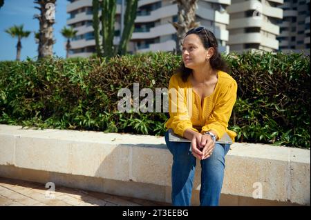 Portrait of a confident pensive authentic pretty woman holding laptop on her knees, sitting on a stone bench outdoor and thoughtfully looking aside, t Stock Photo