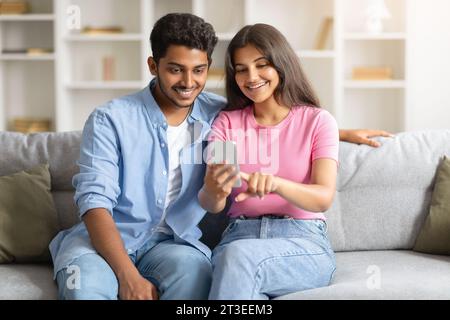 Happy Indian couple on sofa using smartphone, woman pointing at phone screen Stock Photo