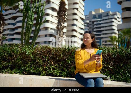 Confident charming curly haired Latin American young adult woman dressed in casual yellow shirt and blue denim, sitting with laptop on her knees, hold Stock Photo