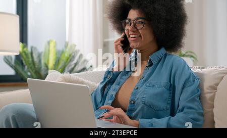 African American girl woman talking mobile phone working with laptop make online order booking food delivery on couch at home lady speaking business Stock Photo