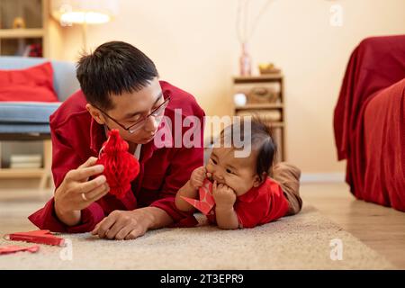 Front view of happy Asian father playing with cute baby boy lying on floor at home and holding red paper decorations for Chinese New Year, copy space Stock Photo